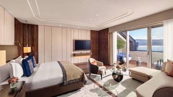 Una habitación de hotel moderna con una cama grande, un televisor, un balcón con vistas al agua y una zona de estar con una mesa de vidrio.
