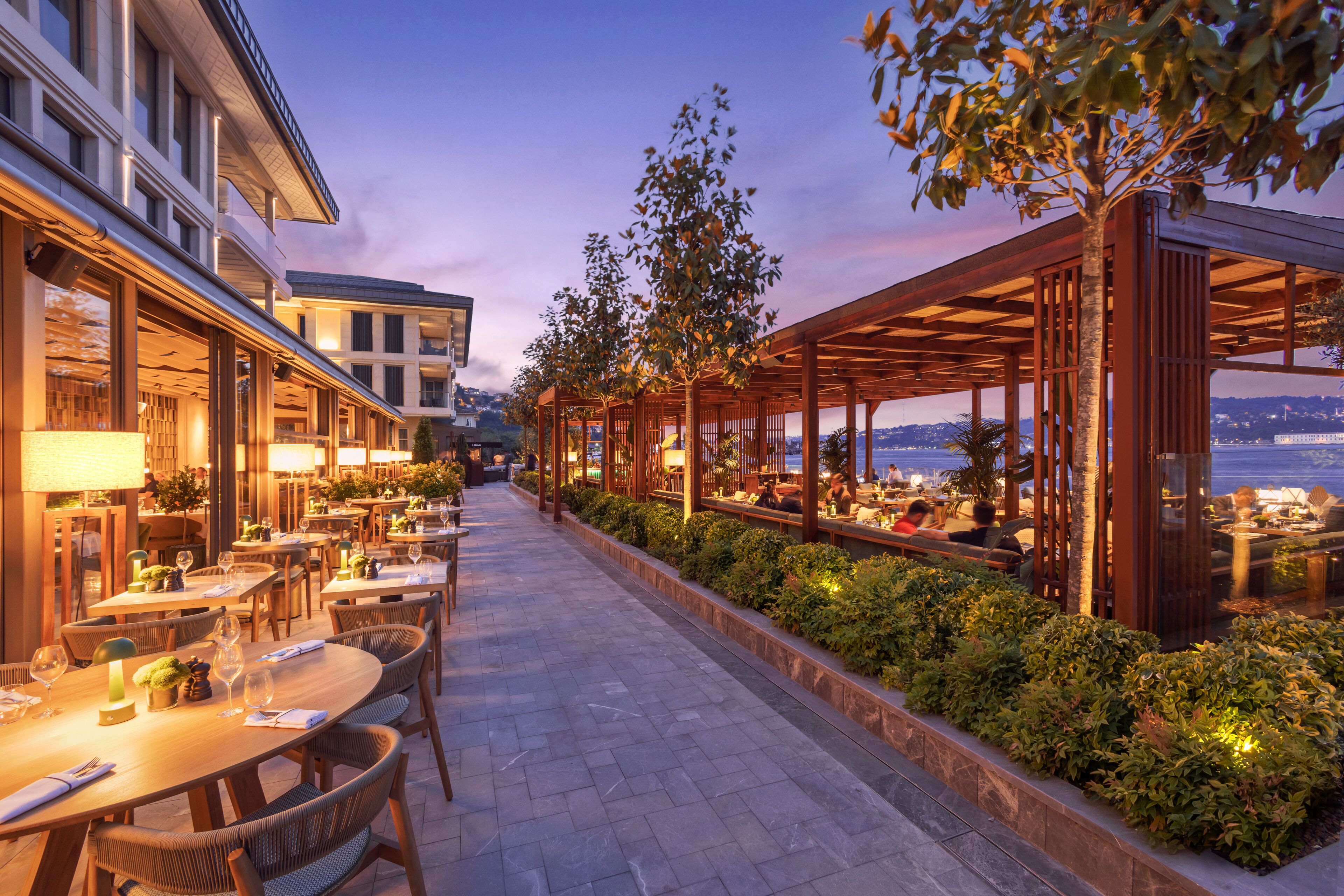 An outdoor dining area with tables and chairs, surrounded by greenery and overlooking a body of water.