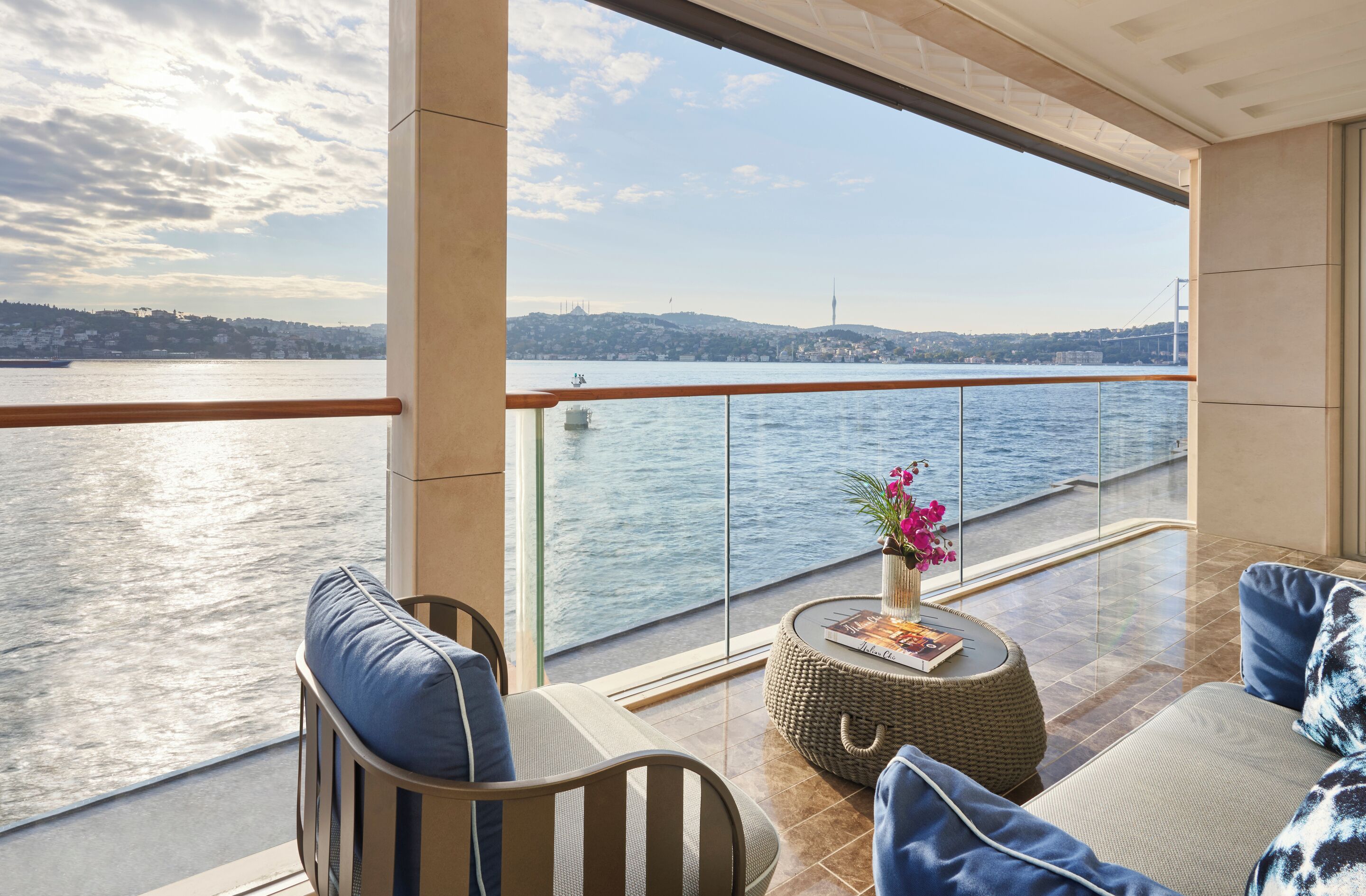 A balcony with a view of a body of water, a bridge, and a distant hill. There are two sofas and a small round table with a vase of flowers.