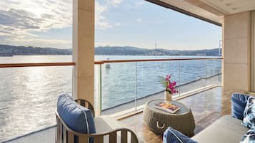 A balcony with a view of a body of water, a bridge, and a distant hill. There are two sofas and a small round table with a vase of flowers.