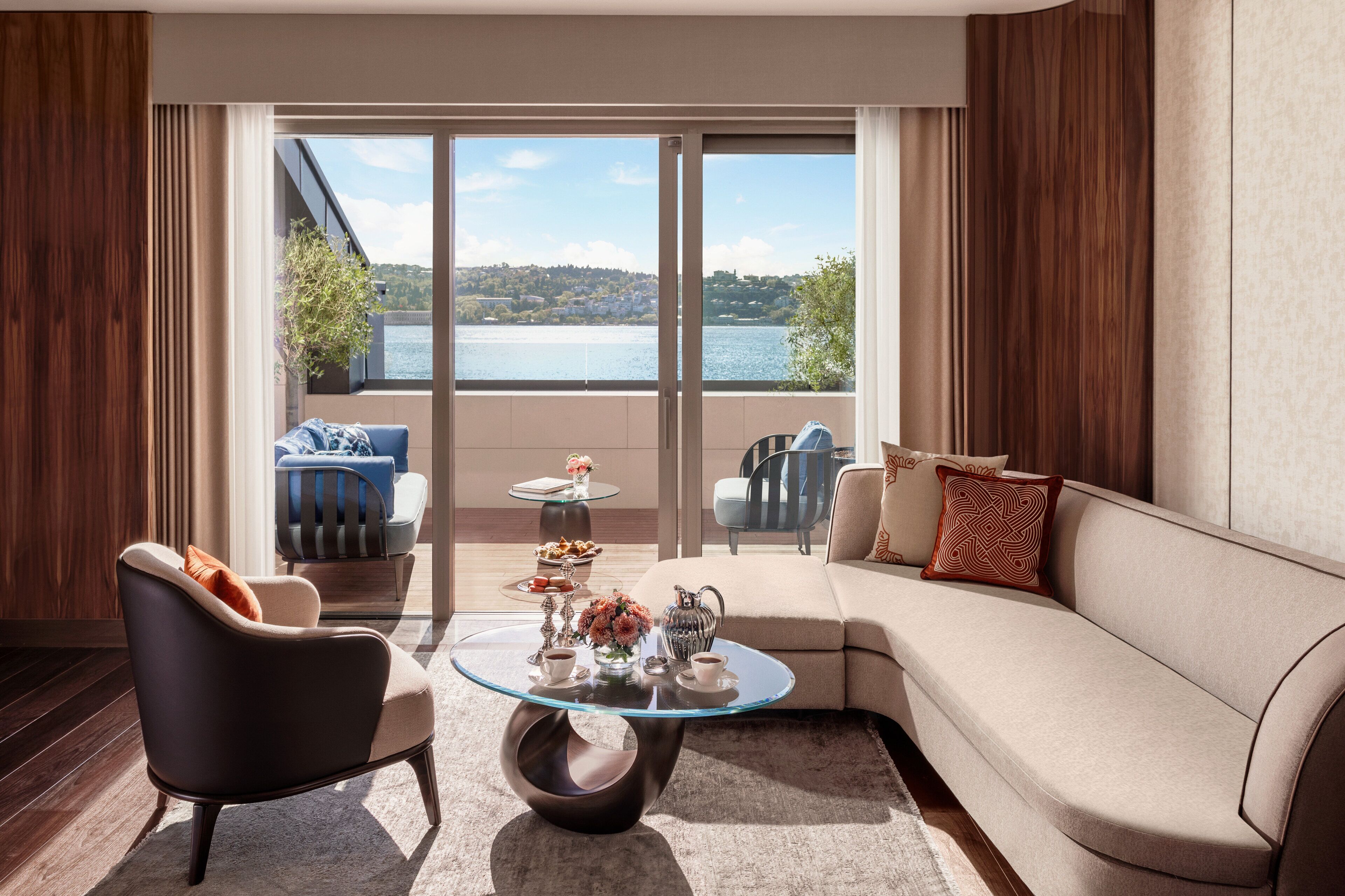 Un salon moderne avec une table basse en verre, un canapé et un fauteuil. De grandes fenêtres offrent une vue sur un lac et des collines.
