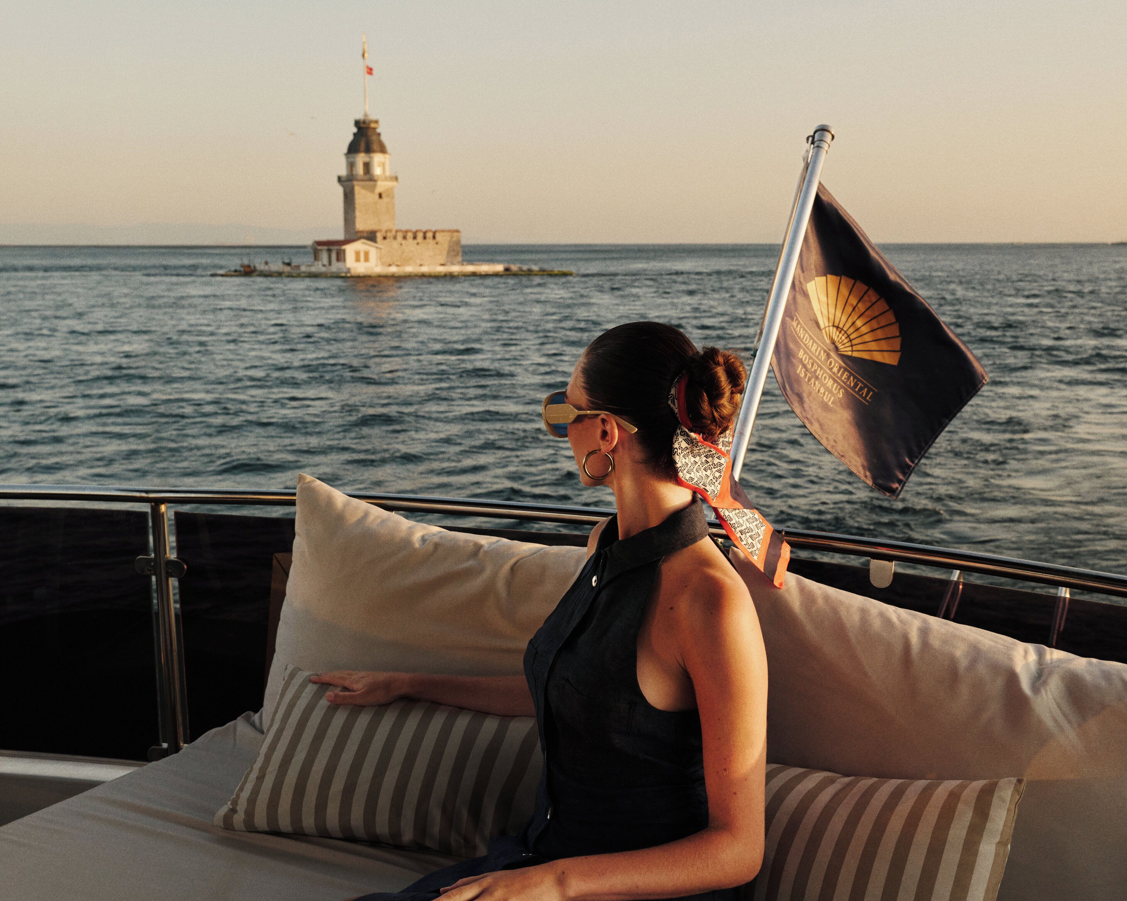 A person on a boat with a flag, a lighthouse in the background, and a body of water.