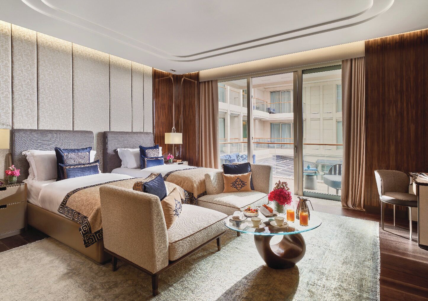 A hotel room with two beds, a sofa, a glass coffee table, and a view of the outside balcony.