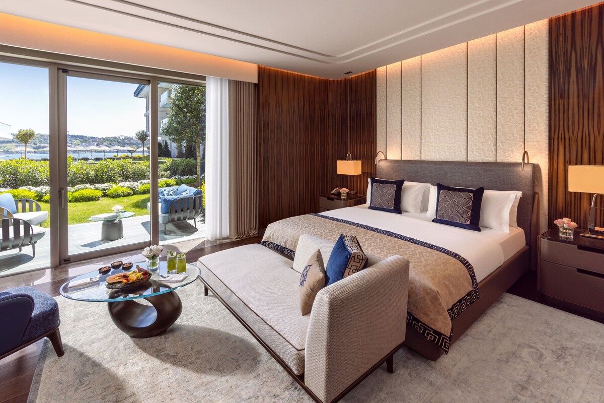A hotel room with a large bed, a glass coffee table, a sofa, and a view of a landscaped garden.
