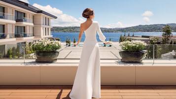 A woman in a white dress standing on a balcony overlooking a body of water with mountains in the distance.