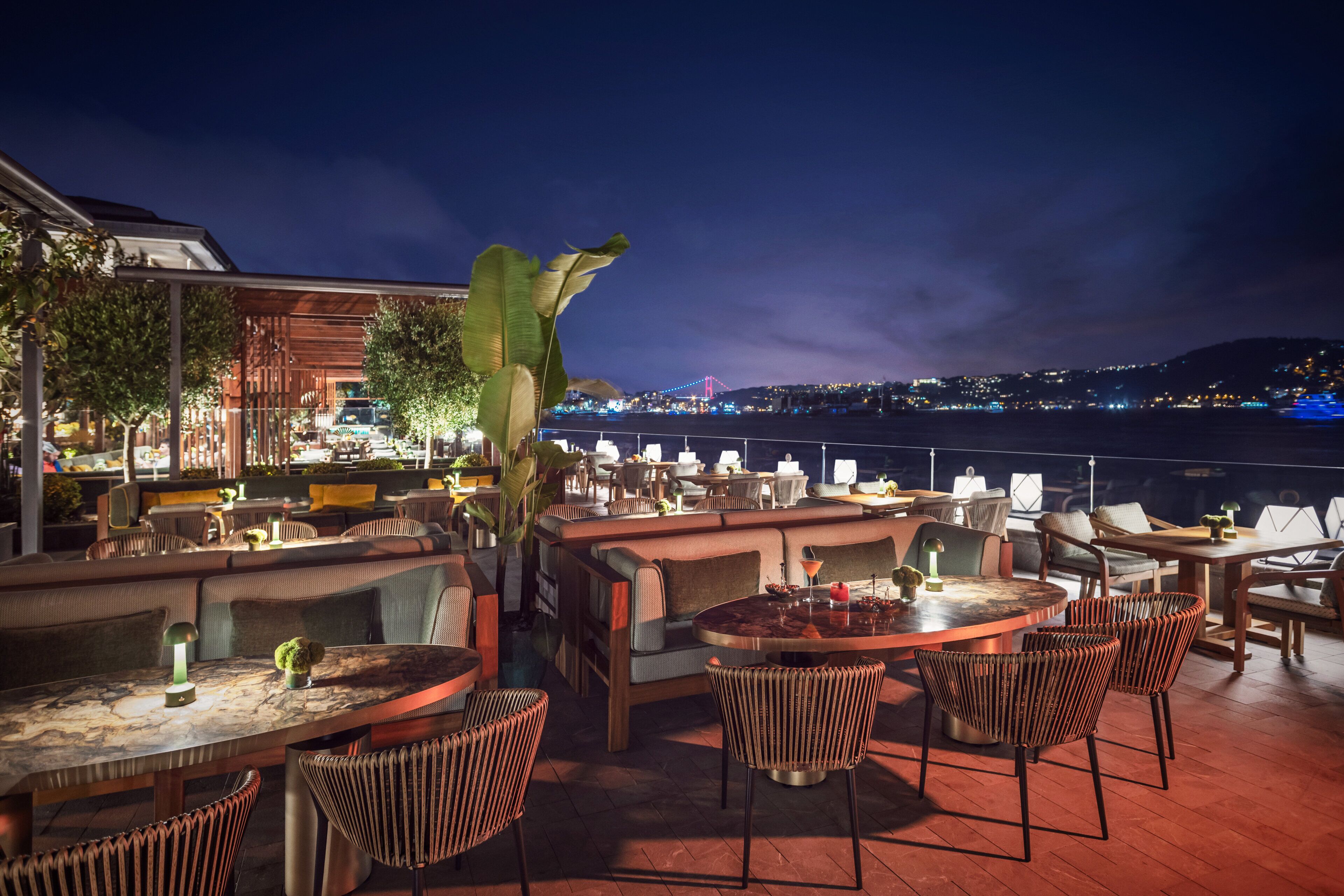 An outdoor dining area with tables and chairs, a view of a bridge, and a cityscape in the background.