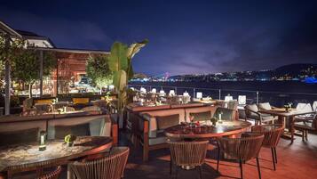An outdoor dining area with tables and chairs, a view of a bridge, and a cityscape in the background.
