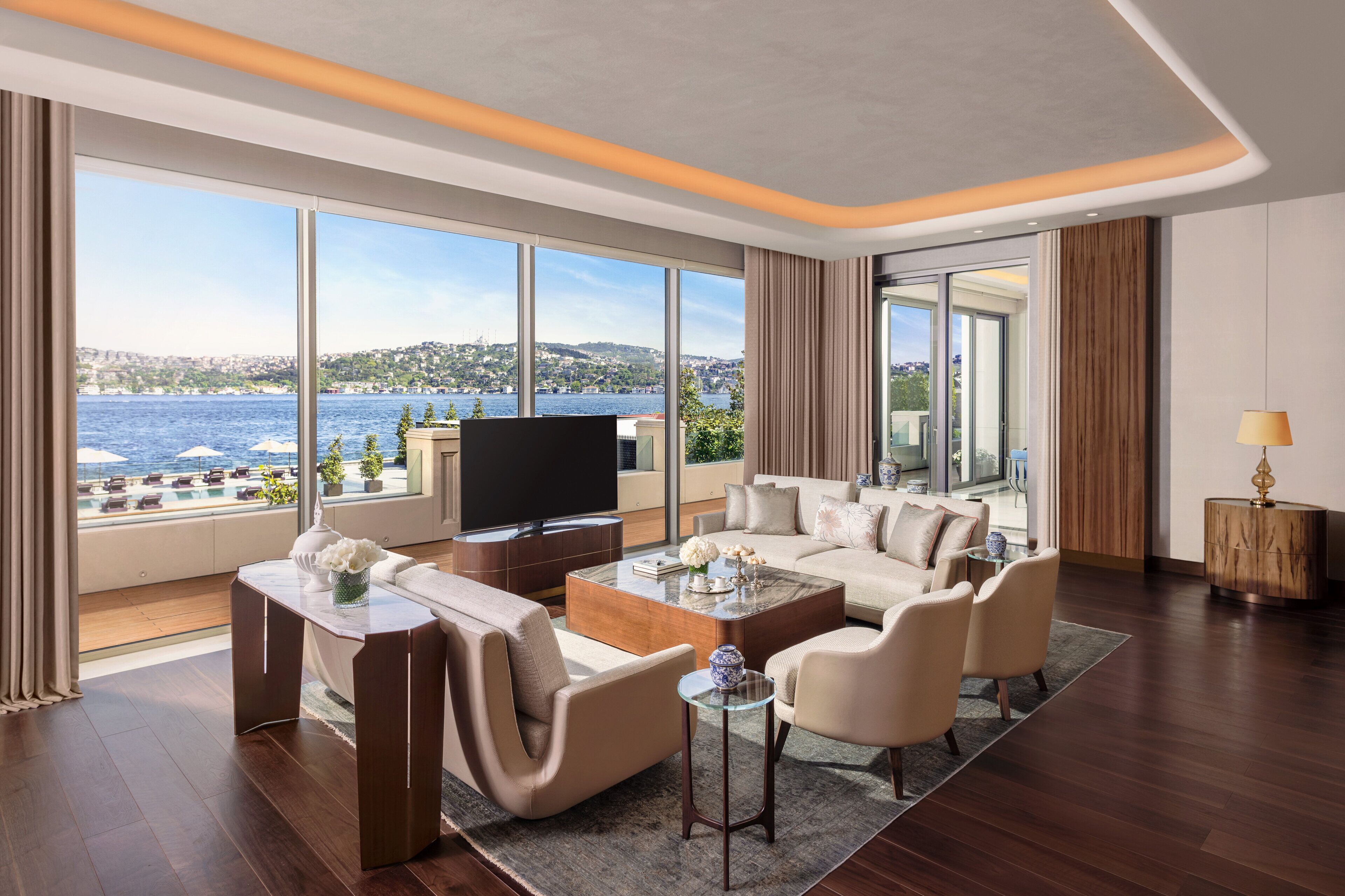 A modern living room with a large TV, comfortable seating, and a view of the sea.