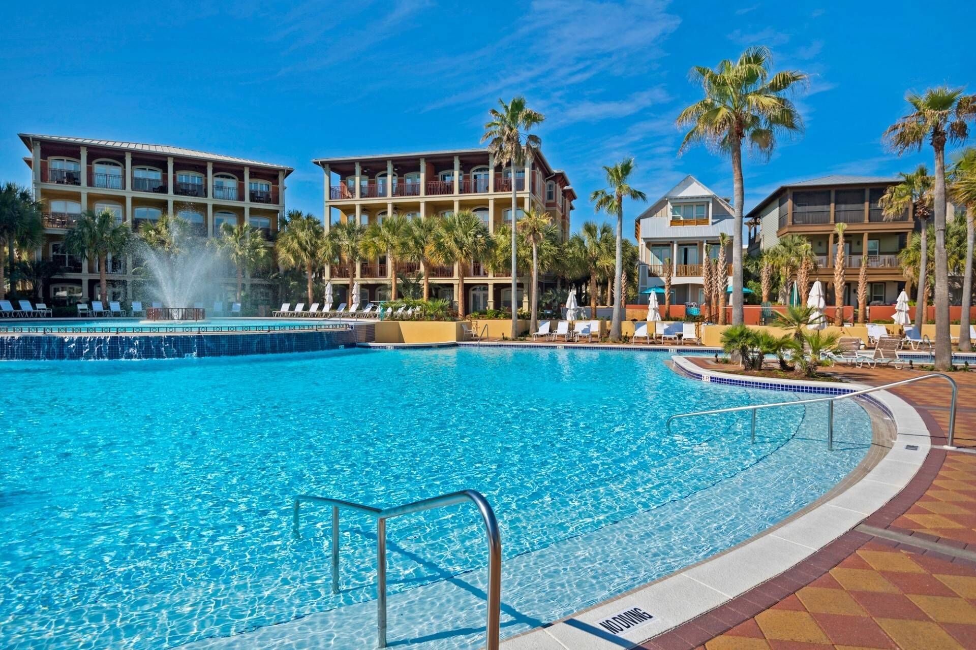 A large outdoor swimming pool with a fountain, surrounded by palm trees and lounge chairs.