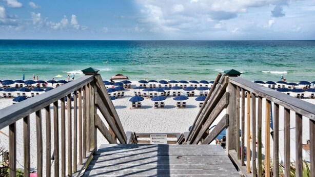 A wooden boardwalk leading to a beach with white sand and blue umbrellas.