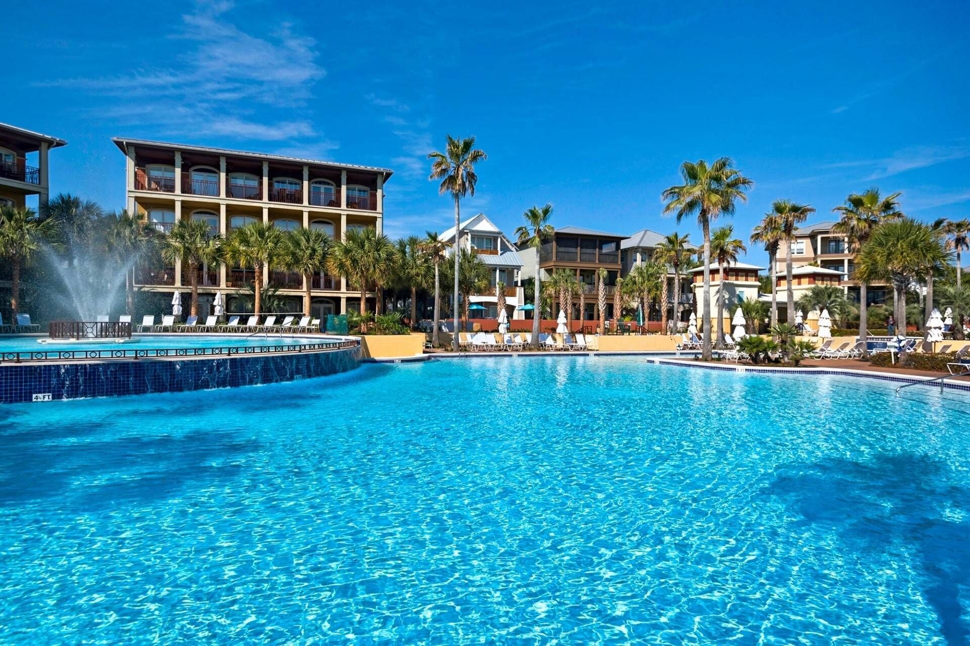 A large, clear swimming pool with lounge chairs and palm trees, surrounded by multi-story hotel buildings.