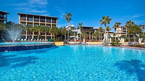 A large, clear swimming pool with lounge chairs and palm trees, surrounded by multi-story hotel buildings.