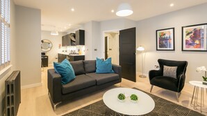 En moderne stue med en grå sofa, en sort lænestol og et rundt sofabord. I baggrunden ses et køkken med et køleskab og en mikrobølgeovn.