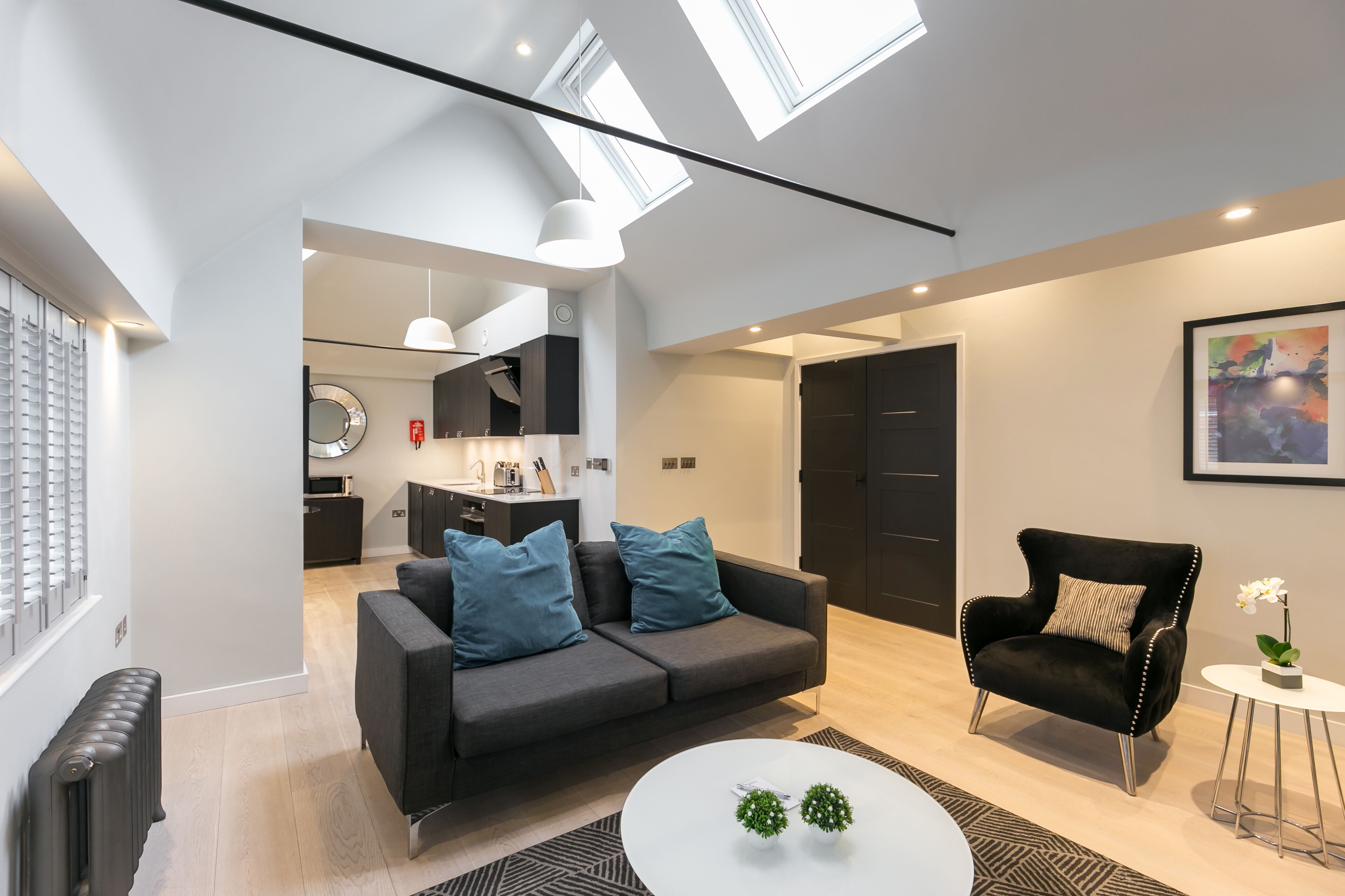 A modern living room with a grey sofa, a black armchair, and a white coffee table. There is a kitchen in the background with a round mirror above the sink.
