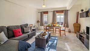A living room with a sofa, coffee table, dining table, chairs, and a TV stand. - Cosy Vacation Home “Casa da Praia” close to the Beach with Wi-Fi (Tavira - Santa Luzia)