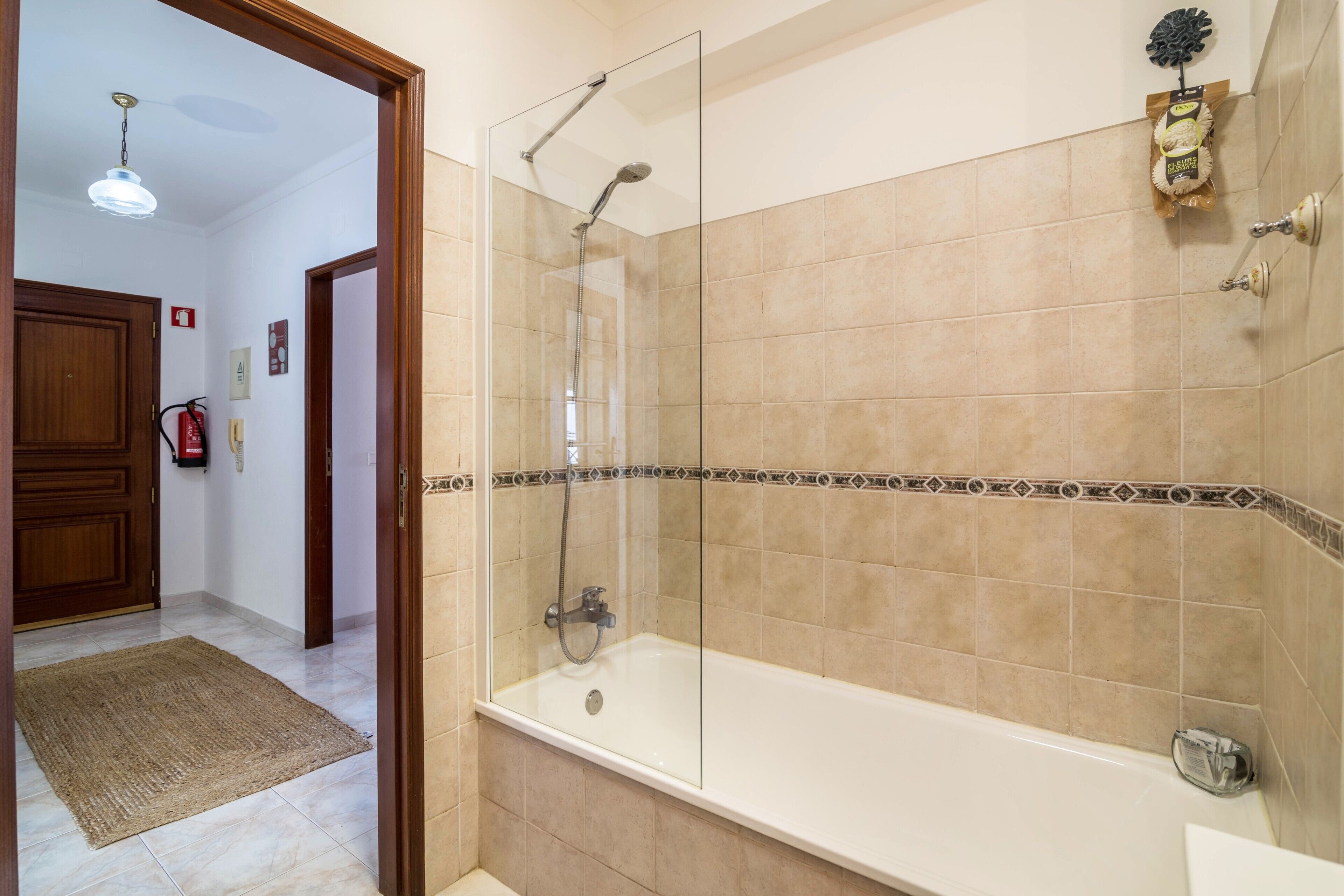 A bathroom with a glass-enclosed shower, a bathtub, beige tiles, and a wooden door.