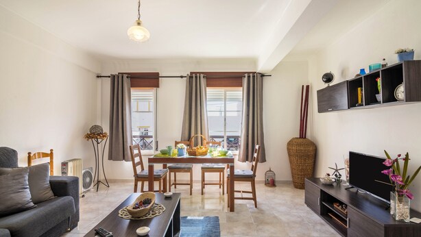 A living room with a sofa, dining table, chairs, TV, and a large vase with flowers. - Cosy Vacation Home “Casa da Praia” close to the Beach with Wi-Fi (Tavira - Santa Luzia)