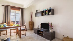 A living room with a dining area, a TV, and a shelf with decorative items. - Cosy Vacation Home “Casa da Praia” close to the Beach with Wi-Fi (Tavira - Santa Luzia)
