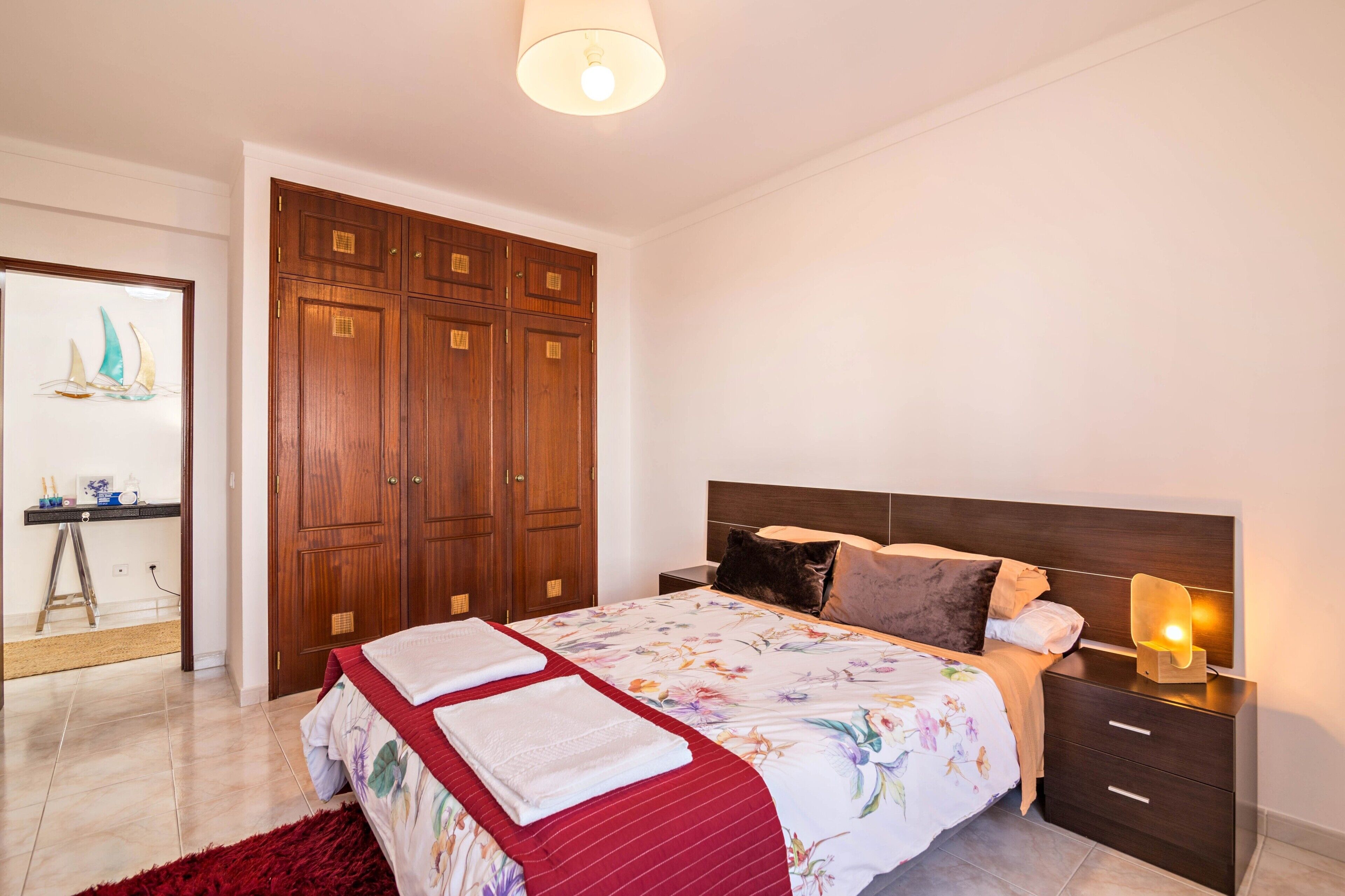 A bedroom with a bed, wooden wardrobe, bedside table, and a lamp.