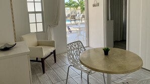 Una habitación con una silla blanca, una mesita con una planta en maceta, una ventana con cortina y vistas a una zona de piscina al aire libre.