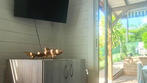 Una habitación con un televisor montado en la pared, un mueble de madera con cajones y vistas a un jardín a través de una puerta de cristal.