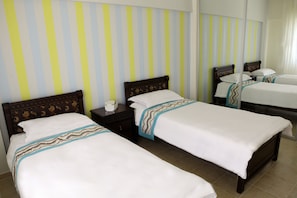 A hotel room with three single beds, each with a wooden headboard and white bedding. There is a nightstand with a lamp and a small table between the beds. The wall has a striped pattern. - Two bed Furnished Apartment in Amman (Amman)
