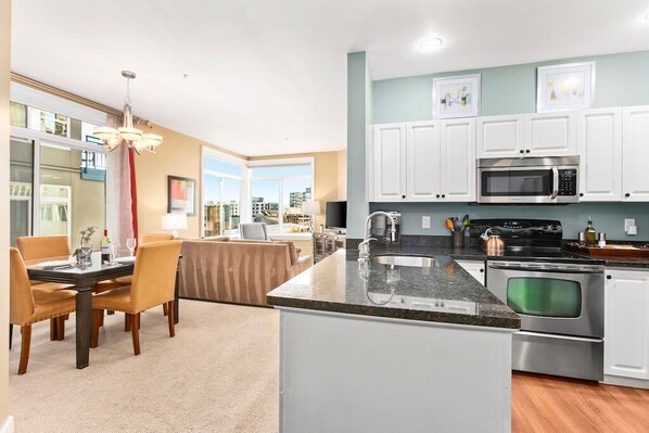 A modern kitchen with white cabinets, a granite countertop, stainless steel appliances, and a dining area with a table and chairs. - Corner Unit in the middle of it all - 99 Walkscore! (Seattle)