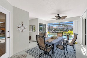 Spiseområde med et bord, stole, ventilator og udsigt til en swimmingpool og bygninger.