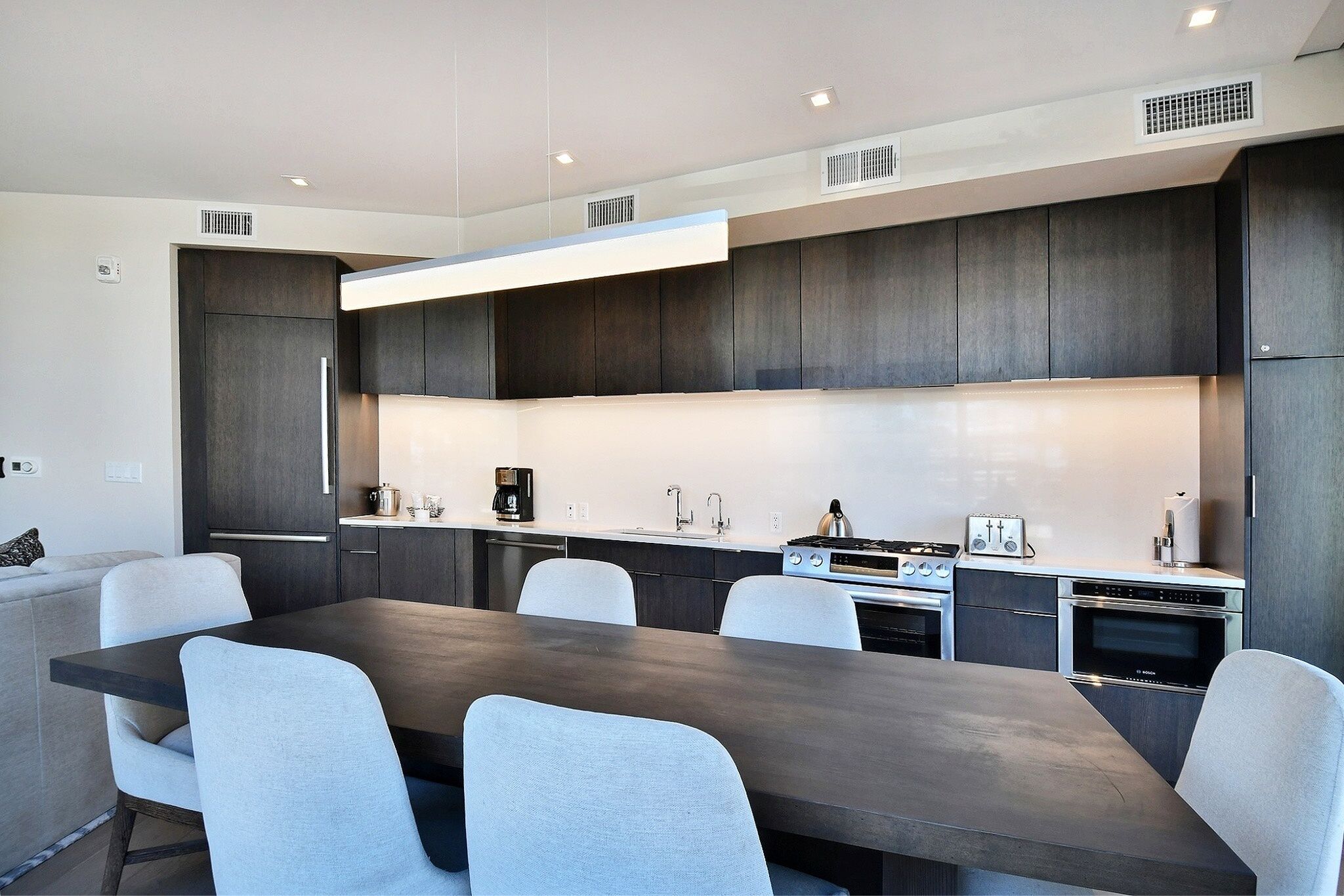 A modern kitchen with dark cabinetry, a central island, and a dining area with chairs.