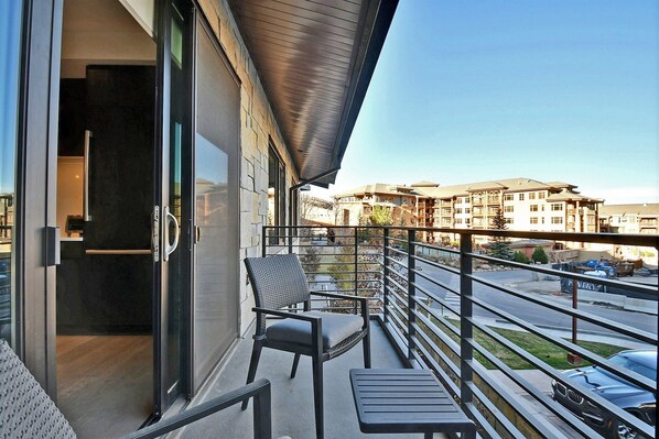 A balcony with a chair and table, a glass door leading to a room, and a view of a building and street in the background.