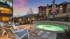 A hot tub surrounded by stone patio, outdoor seating, and a view of a multi-story building with balconies. - Luxury 2 BR in Canyons Village - Walk to Slopes by RedAwning (Park City)