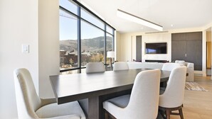 A modern dining area with a large table and chairs, a flat-screen TV mounted on the wall, and a view of a mountainous landscape through large windows. - Luxury 2 BR in Canyons Village - Walk to Slopes by RedAwning (Park City)