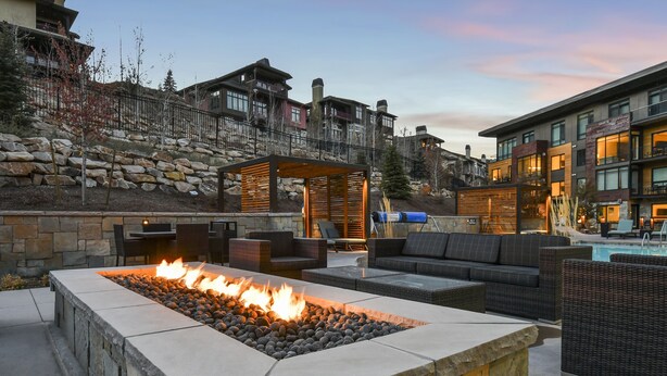 A stone fire pit with seating, a pool, and a stone wall with a wooden pergola. - New 1 BR Residence in Canyons Village- Ski in/out by RedAwning (Park City)