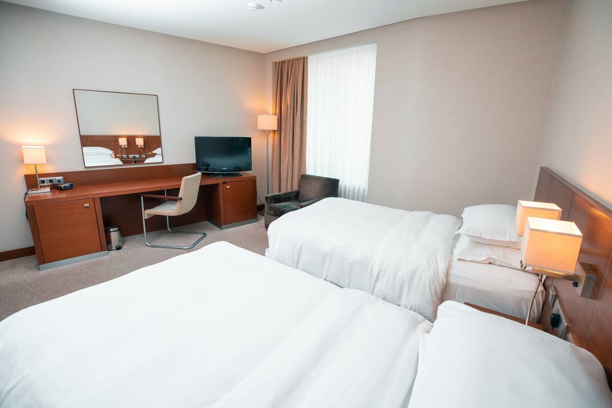 A hotel room with a desk, two beds, a TV, and a large window with curtains.