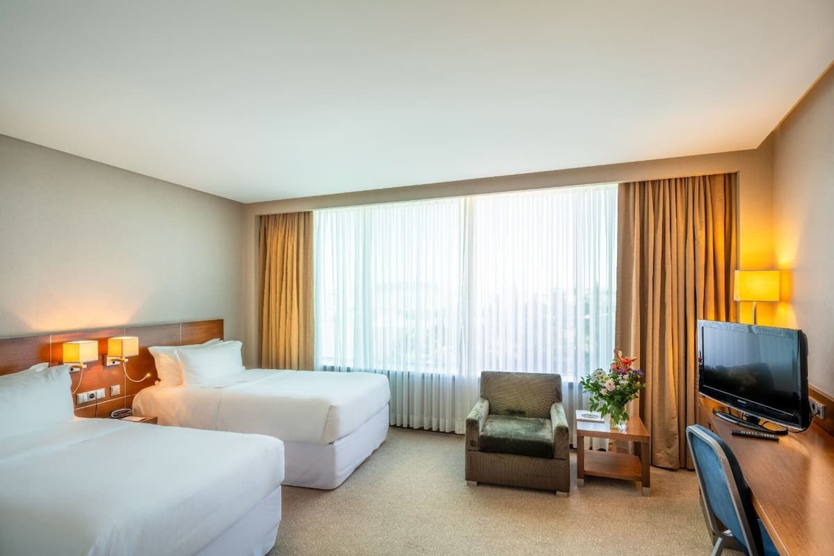 A hotel room with two beds, a chair, a desk with a TV, and a window with curtains.