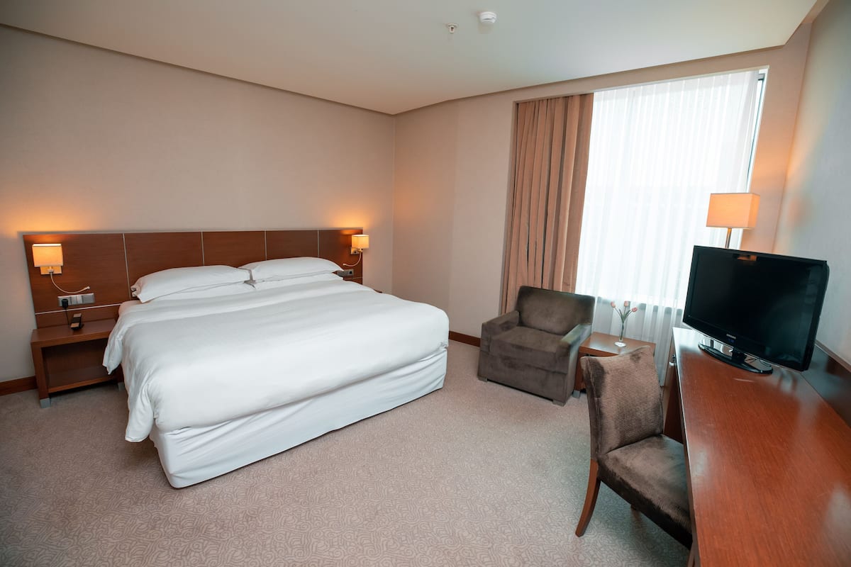 A hotel room with a large bed, a desk with a TV, a chair, and a nightstand with a lamp.