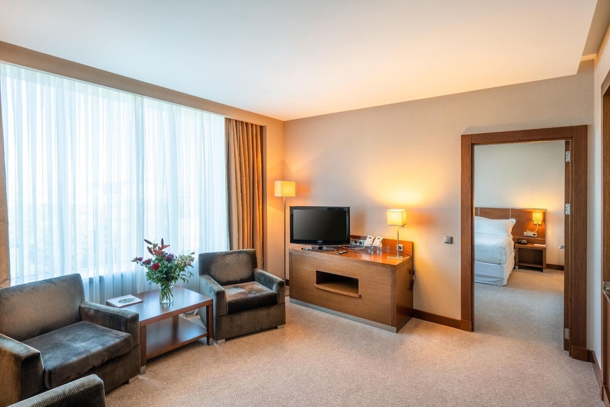 A hotel room with a sofa, armchair, TV, desk, lamp, and a bed in the background.