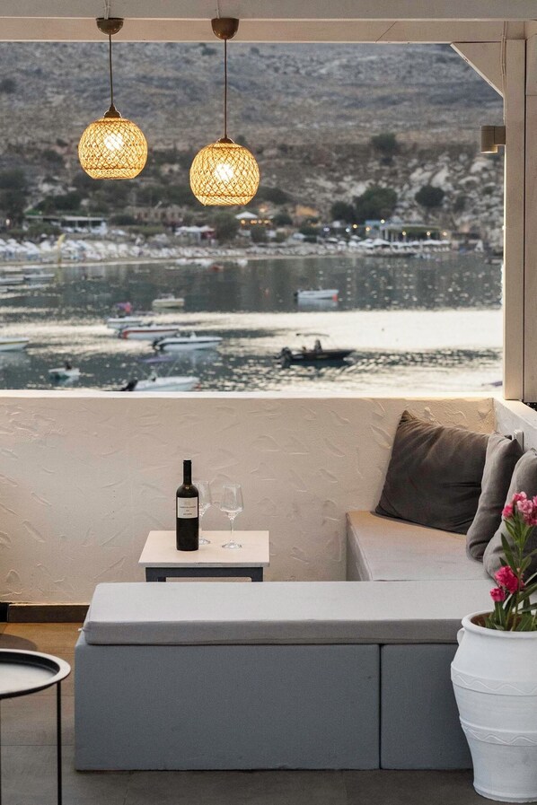 Un balcon avec un canapé, une table avec une bouteille et des verres, des lampes en osier suspendues, et une vue sur une étendue d’eau et des bâtiments.