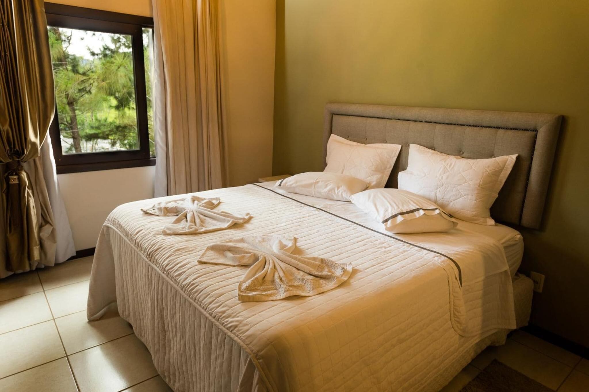 Una cama bien hecha con colcha acolchada y almohadas, una ventana con cortinas y vistas al exterior.