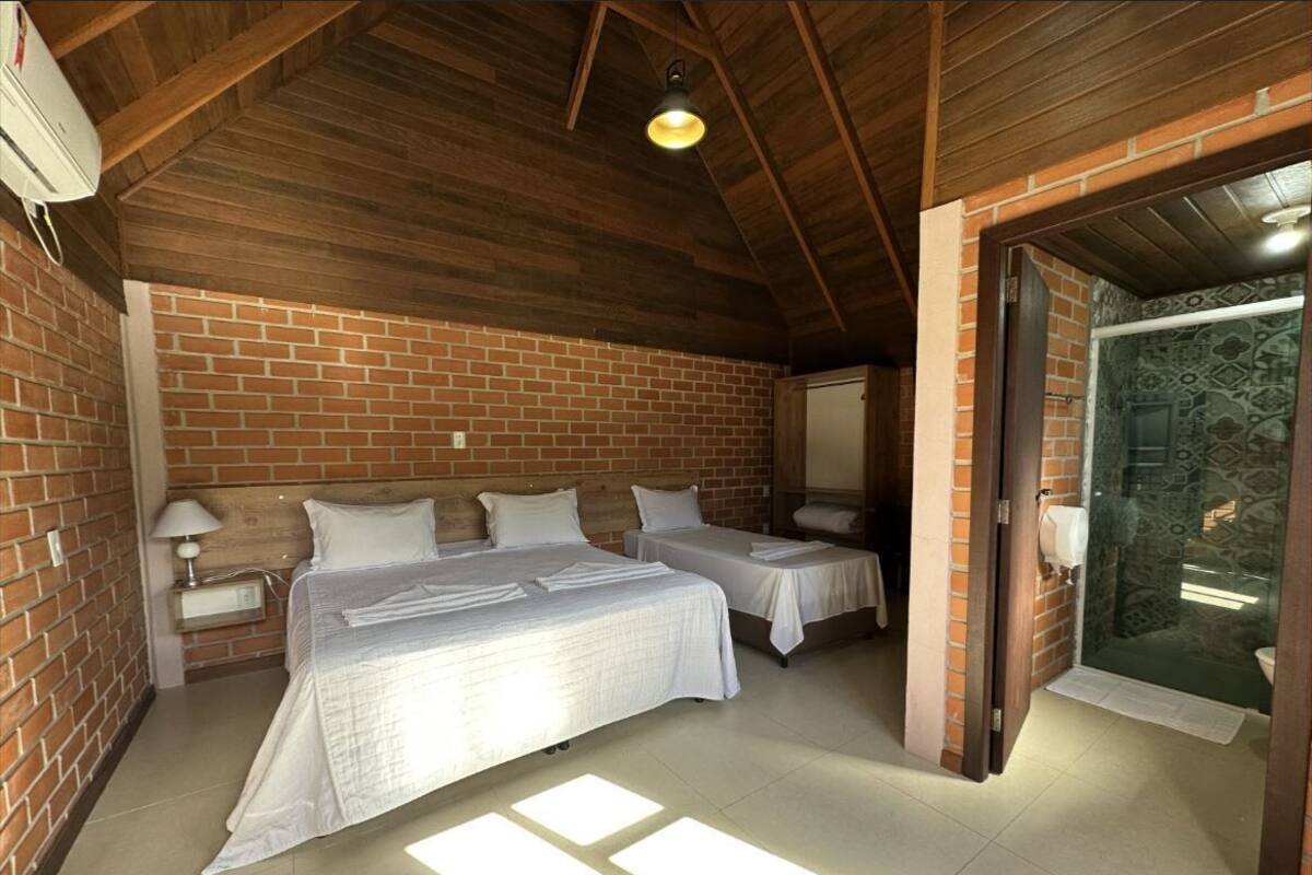 A room with two beds, a brick wall, a wooden ceiling, and a glass door leading to a bathroom.