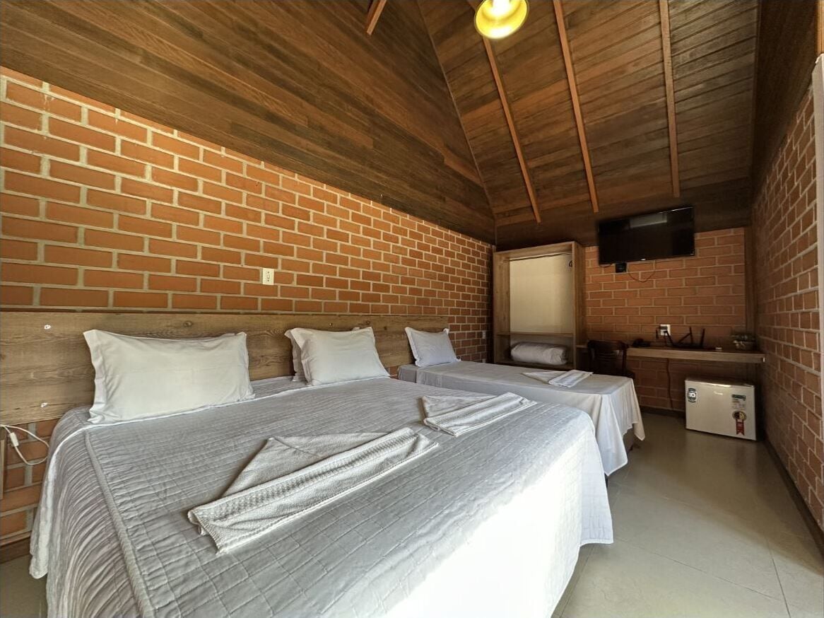 Une chambre avec deux lits, un plafond en bois, des murs en briques, une petite kitchenette et un téléviseur fixé au mur.