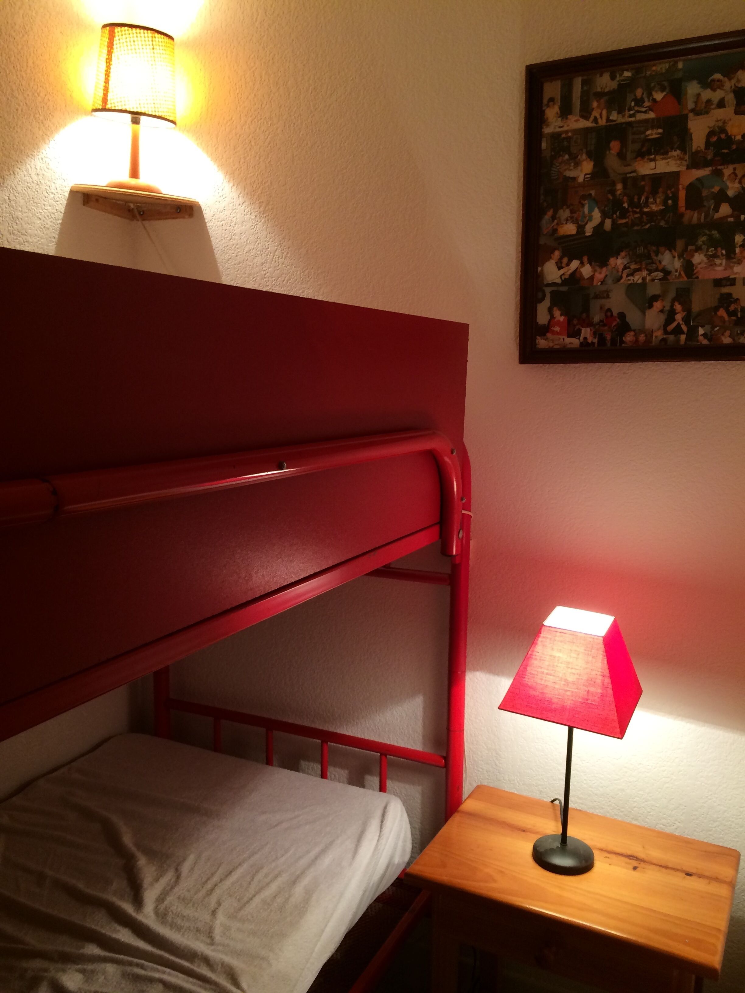 Ein Etagenbett mit rotem Kopfteil und einem Einzelbett darunter. Ein kleiner Nachttisch aus Holz mit einer roten Lampe steht daneben.