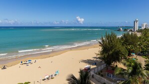 A beach with clear turquoise waters, a sandy shore, and a few umbrellas. There are palm trees and a few buildings in the background.
