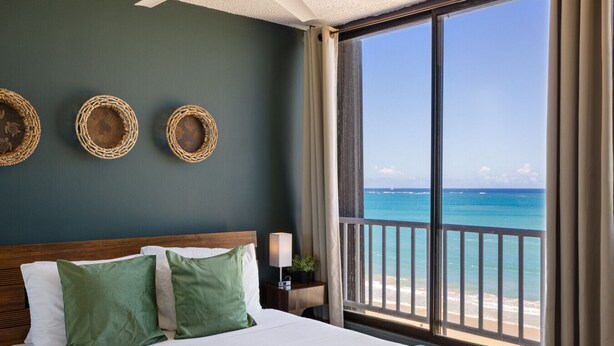 A hotel room with a large window overlooking a beach, a bed with green pillows, and three woven wall decorations.