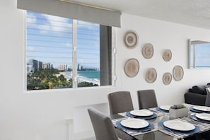 A dining area with a city view, a table set with plates and cutlery, and woven wall decor. - Flamenco Beach 2BDR Oceanfront Pool Parking (Isla Verde)