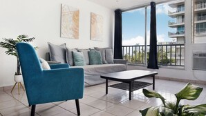 A modern living room with a sofa, a blue armchair, and a coffee table. There are two abstract paintings on the wall, a ceiling fan, and a view of a building through large windows. - Luna Llena Beach 1BDR Oceanview Pool (Isla Verde)