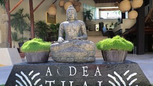 Une statue de Bouddha, placée sur un piédestal, porte l’inscription « A DEA THAI » et est entourée de plantes en pot.