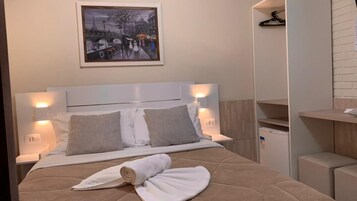 Et hotelværelse med seng, to puder, et håndklæde, et natbord, en lampe, et lille køleskab, et billede på væggen og en træhylde.