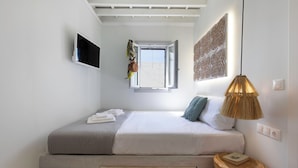 Ein Schlafzimmer mit Bett, Fernseher, einem Fenster mit Kleidern, einer Lampe und einem kleinen Holztisch.