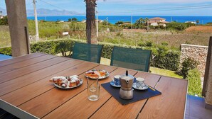 A wooden table with plates of food, a coffee pot, and cups, set against a scenic view of the sea and hills. - VILLA CIURIDDA (Alcamo)